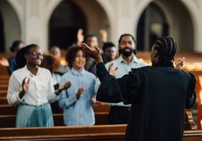 African american preacher is leading her congregation in prayer, with their hands raised in worship. They are all gathered together in the church pews